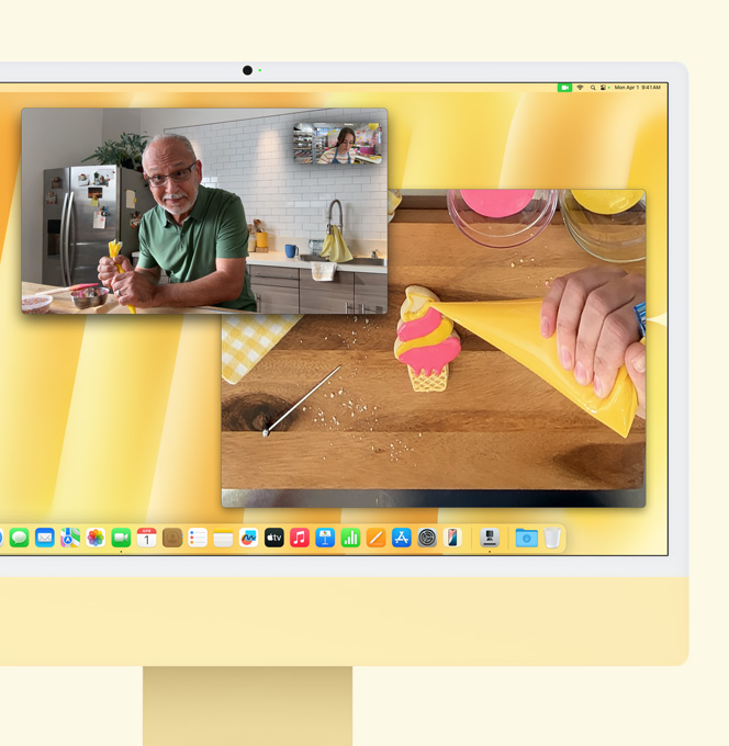 iMac Kuning. Demo Desk View. Layar menampilkan pembuat kue sedang dalam panggilan video dengan seseorang. Satu layar menampilkan wajah seorang pria dan pembuat kue tersebut. Layar lainnya menampilkan tangan pembuat kue yang sedang menghias kue