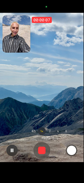 Una persona che riprende se stessa con la fotocamera frontale e contemporaneamente riprende un paesaggio montano con la fotocamera posteriore per mettere in evidenza la funzione Acquisizione doppia