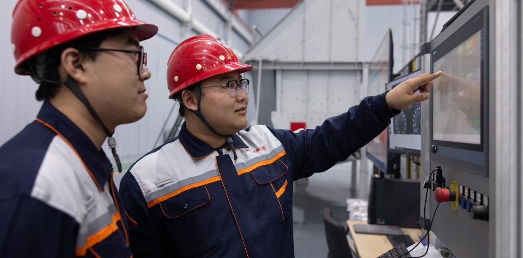 Two employees wearing safety hats and uniforms look at digital screen with control handles at the bottom