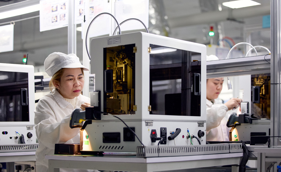 Two employees in work smocks and hats stand at their work stations operating Apple Watch assembly machines
